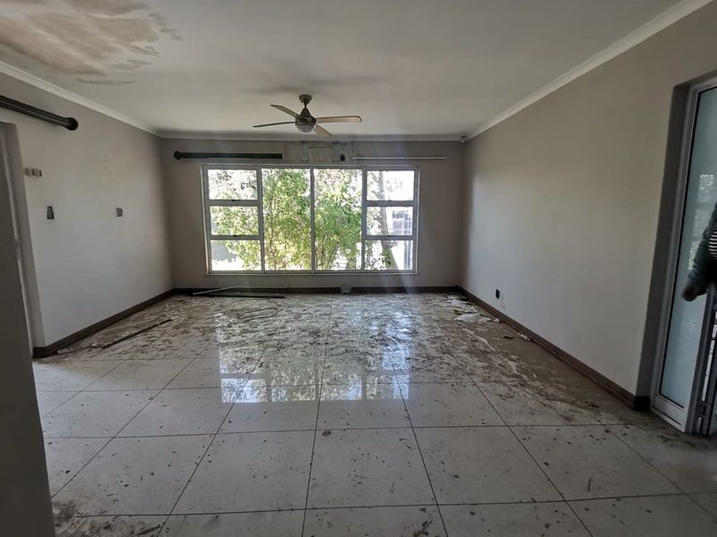 An empty living room featuring tiled floors, a ceiling fan, and large windows, currently showing signs of water damage and requiring significant maintenance.