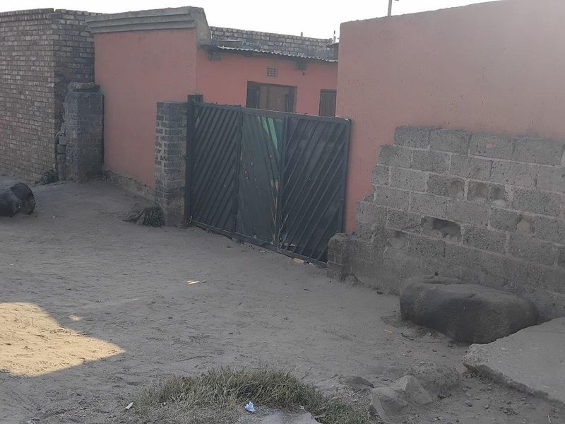 Street view of a residential property featuring a boundary wall and metal gate access from an unpaved road.