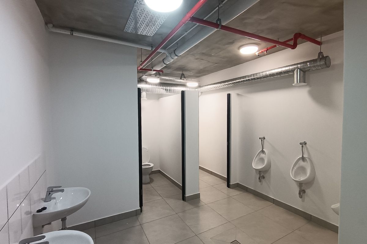 Modern industrial-style communal bathroom featuring exposed ceiling pipes, tiled flooring, multiple urinals, and private toilet cubicles.