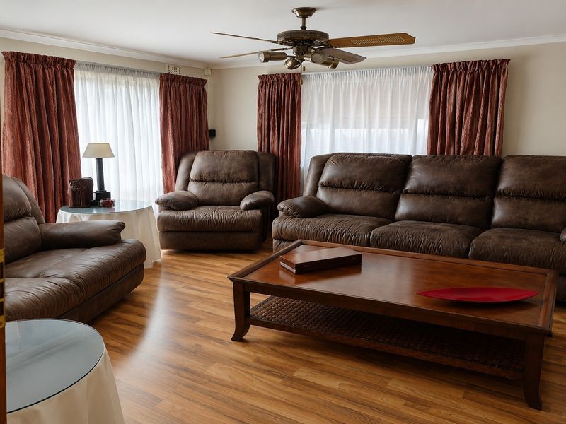A comfortable living room featuring laminate wood flooring, a central ceiling fan, and large windows dressed with curtains.