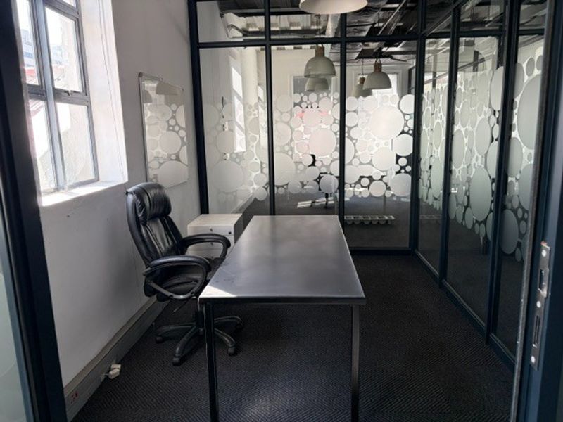 A modern private office space featuring glass partition walls with decorative frosted film, carpeted flooring, and an exposed ceiling with pendant lighting.