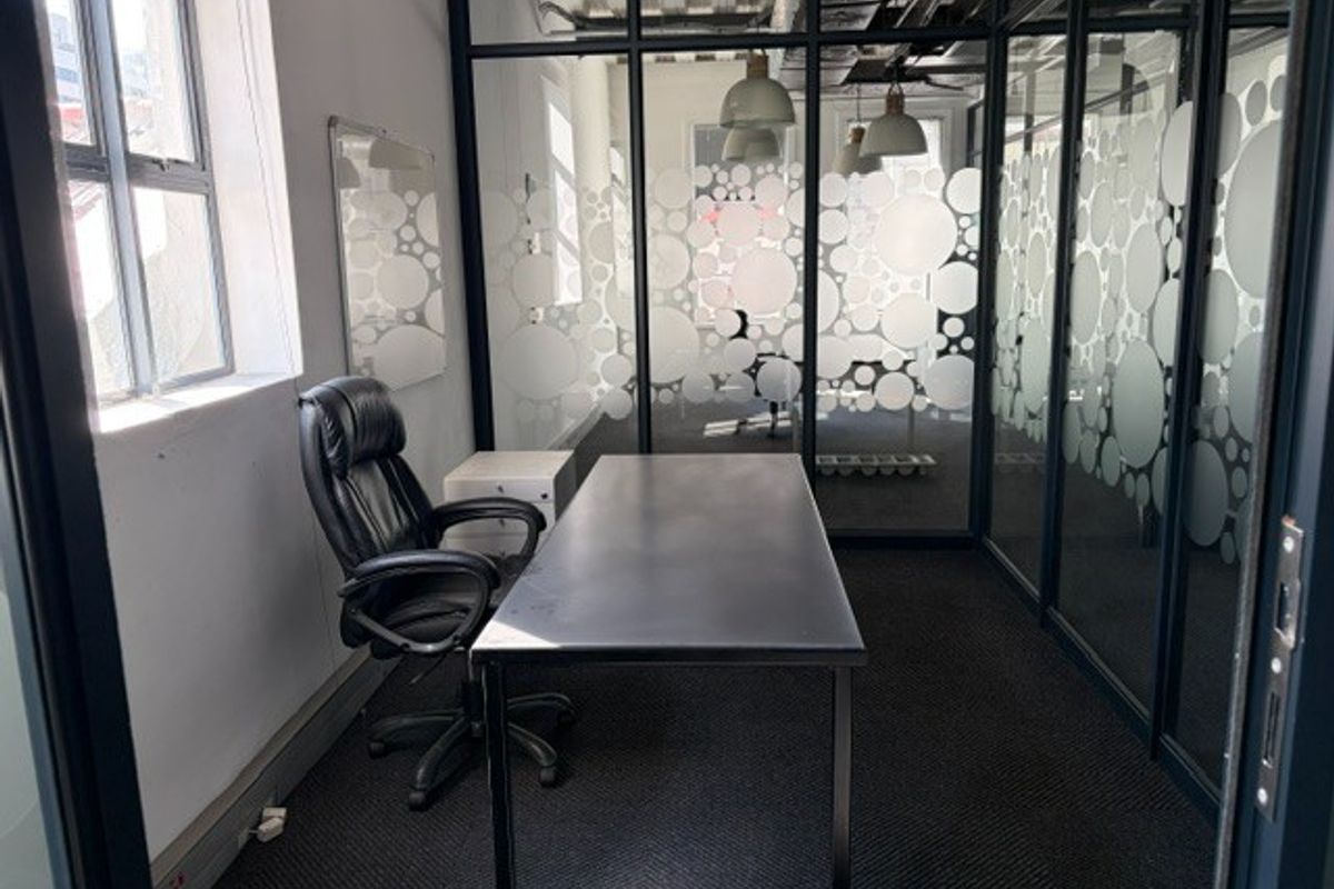 A modern private office space featuring glass partition walls with decorative frosted film, carpeted flooring, and an exposed ceiling with pendant lighting.