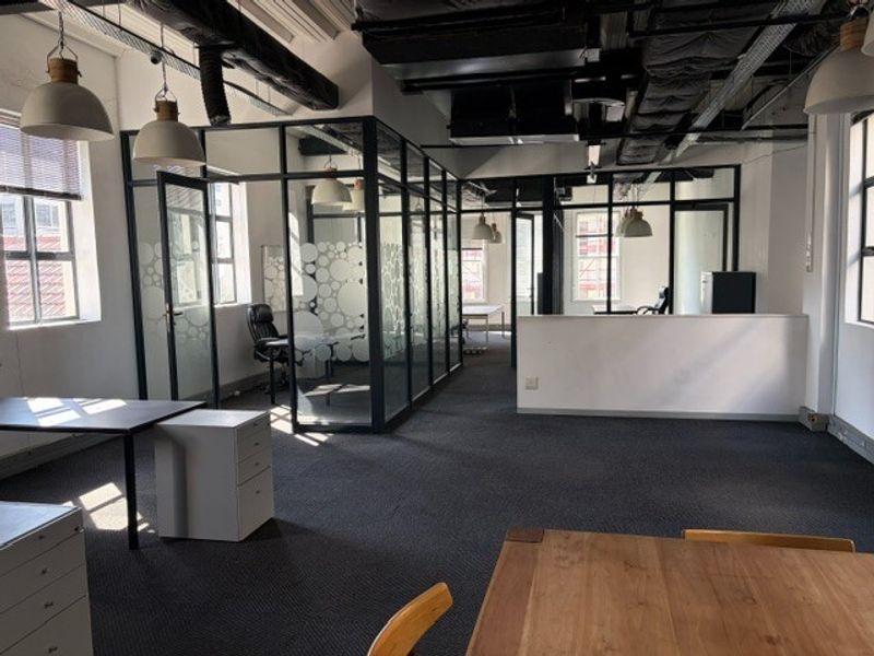 Modern open-plan office space featuring glass-partitioned meeting rooms, exposed industrial-style ceiling ductwork, and neutral carpet flooring.