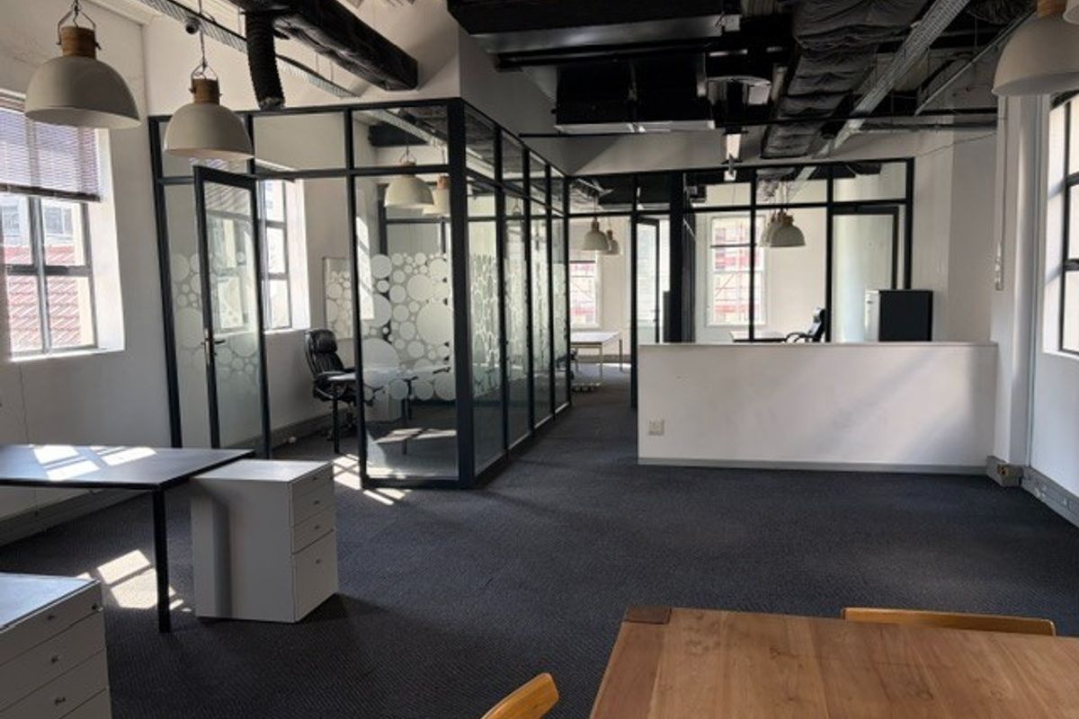 Modern open-plan office space featuring glass-partitioned meeting rooms, exposed industrial-style ceiling ductwork, and neutral carpet flooring.