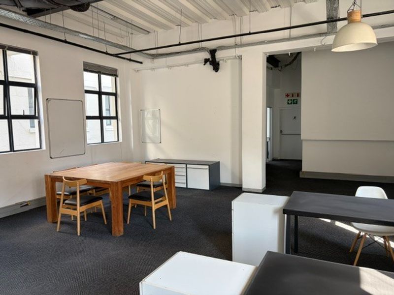 A spacious office area featuring an industrial-style exposed ceiling, large windows for natural light, and carpeted flooring.
