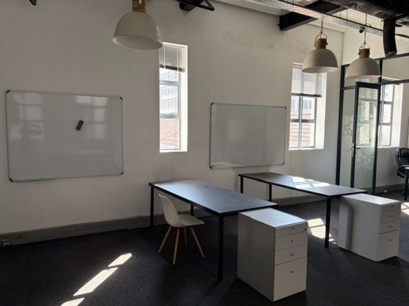 A functional office space featuring exposed ceilings, pendant lighting, wall-mounted whiteboards, and large windows providing natural light.