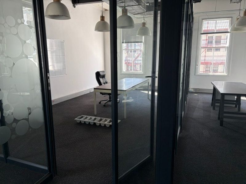 A modern office space featuring glass partition walls, carpeted flooring, and large windows providing natural light.
