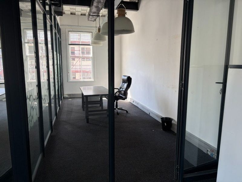 A private office space featuring glass partition walls, exposed industrial-style ceiling, pendant lighting, and dark carpeting.