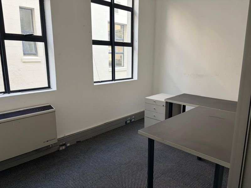 A functional office space featuring large black-framed windows, carpeted flooring, and a built-in HVAC unit, providing a practical workspace environment.