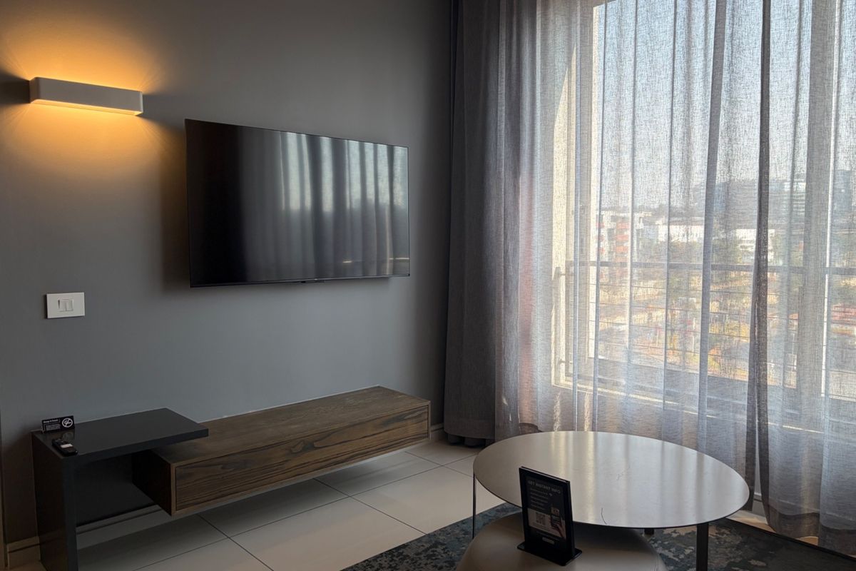 Modern living room featuring a wall-mounted television, floating media console, warm wall lighting, and large windows with sheer curtains.