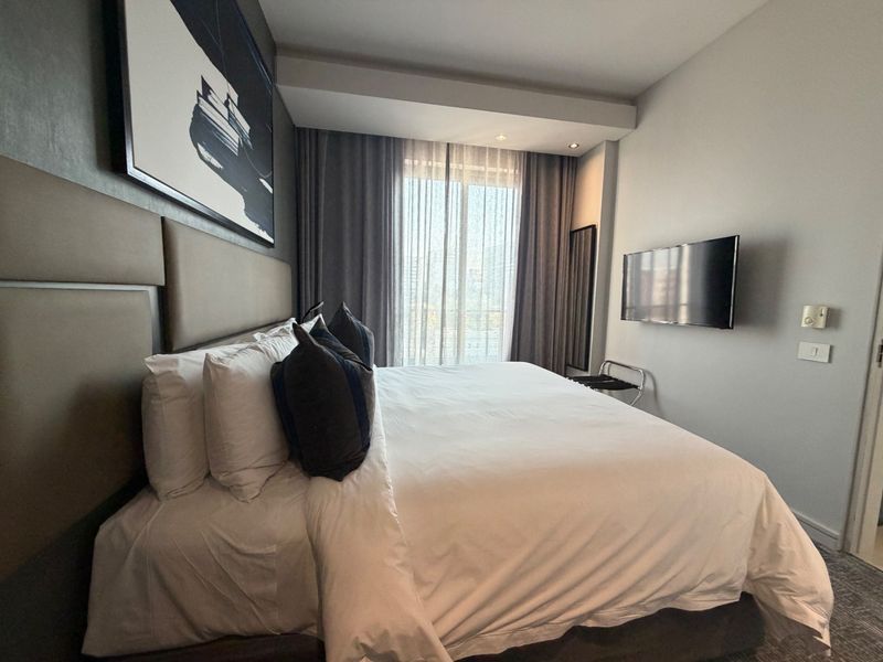A well-appointed bedroom featuring a large upholstered headboard, recessed ceiling lighting, and a wall-mounted television with access to natural light through floor-to-ceiling curtains.