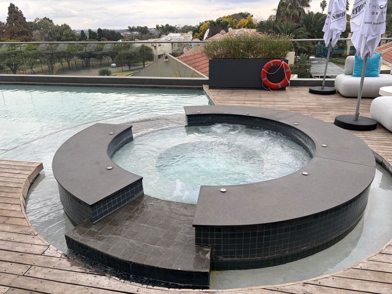 A well-maintained rooftop pool area featuring an integrated spa, timber decking, and a glass balustrade with panoramic views.