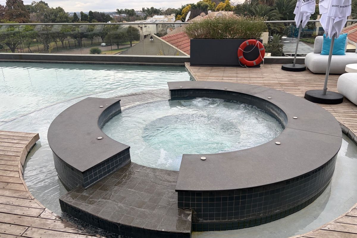 A well-maintained rooftop pool area featuring an integrated spa, timber decking, and a glass balustrade with panoramic views.