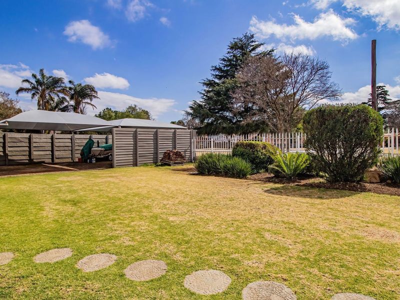 A well-maintained rear garden featuring a lawn, landscaped beds, a paved stepping stone path, and a covered carport structure with boundary fencing.