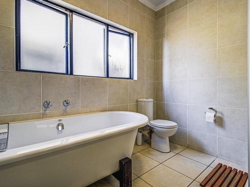 A functional bathroom featuring a classic freestanding bathtub, a standard toilet, and neutral wall and floor tiling with natural light from a frosted window.