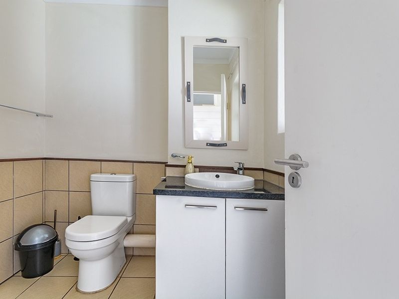 A compact bathroom featuring a white vanity with a basin, a wall-mounted mirror, a toilet, and neutral-toned wall and floor tiling.