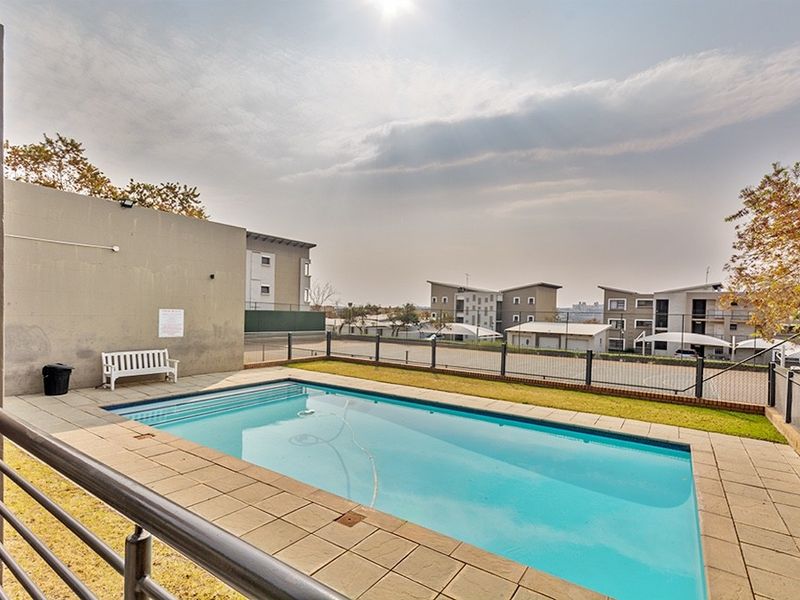 Communal swimming pool area featuring a paved deck, perimeter fencing, and a retaining wall within an apartment complex setting.