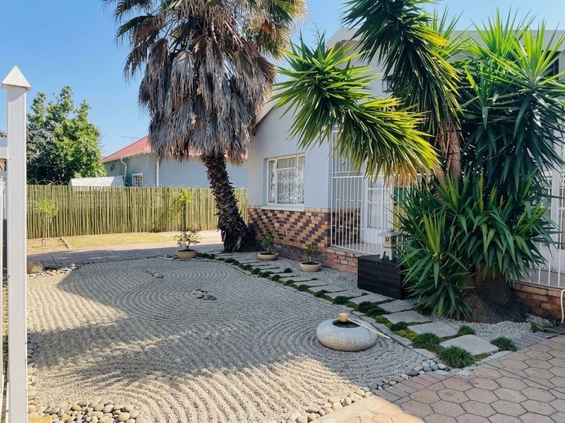 A well-maintained zen-style garden featuring a raked gravel area, stone water feature, paved pathways, and mature palm trees adjacent to the house exterior.