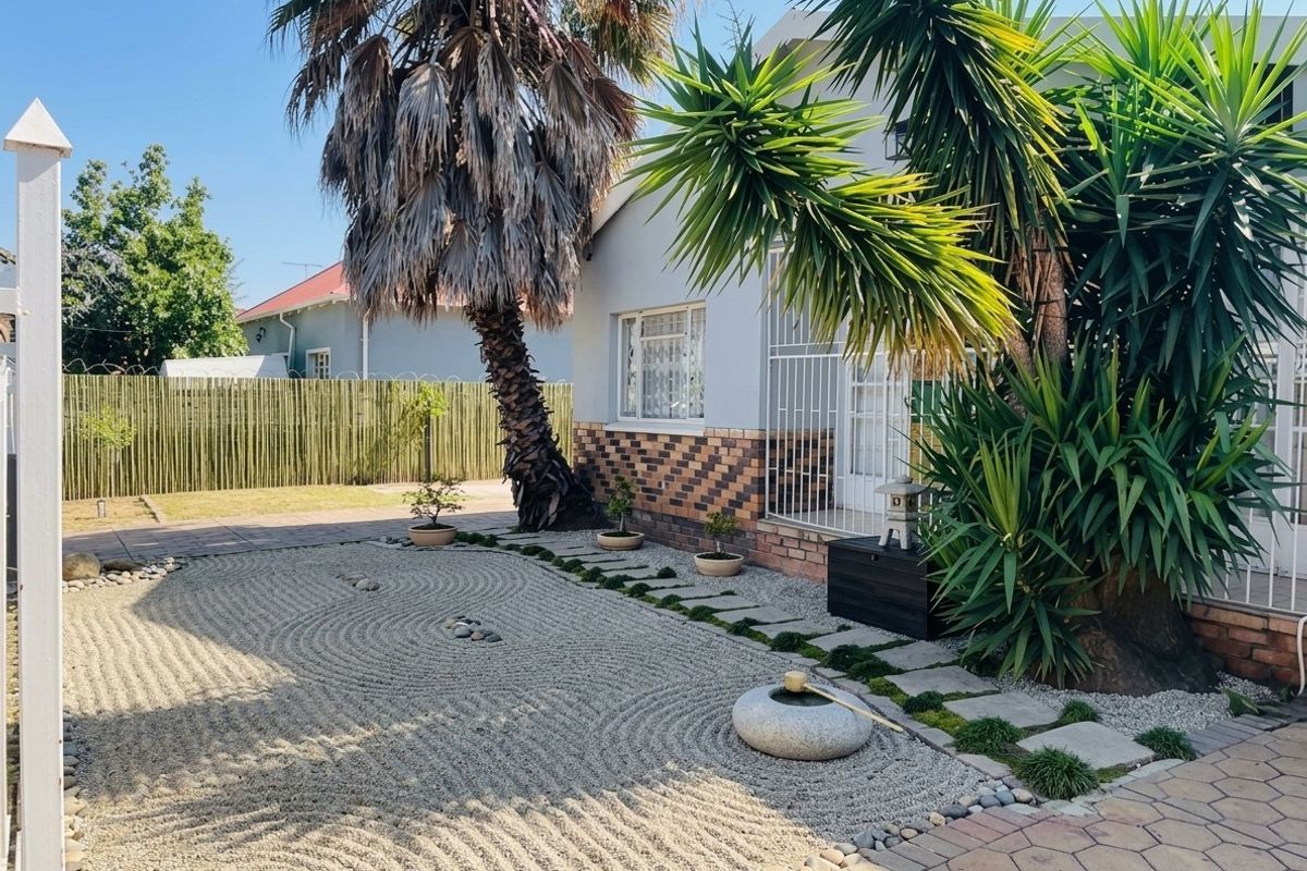 A well-maintained zen-style garden featuring a raked gravel area, stone water feature, paved pathways, and mature palm trees adjacent to the house exterior.