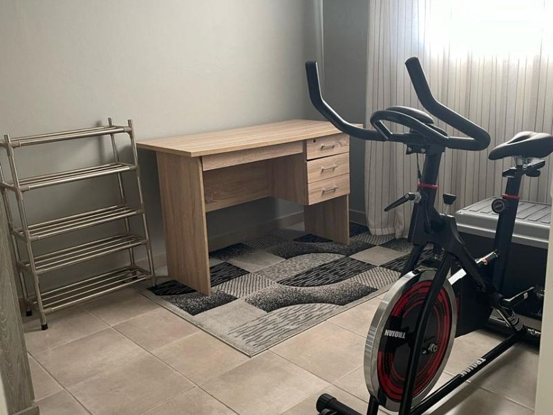 A functional bedroom space featuring neutral tiled flooring and natural light, currently utilized as a home office and fitness area.