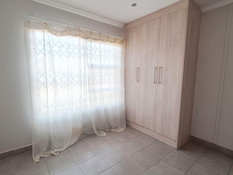 A bright, empty bedroom featuring neutral tiled flooring, built-in wooden cupboards, and a window fitted with a security gate.