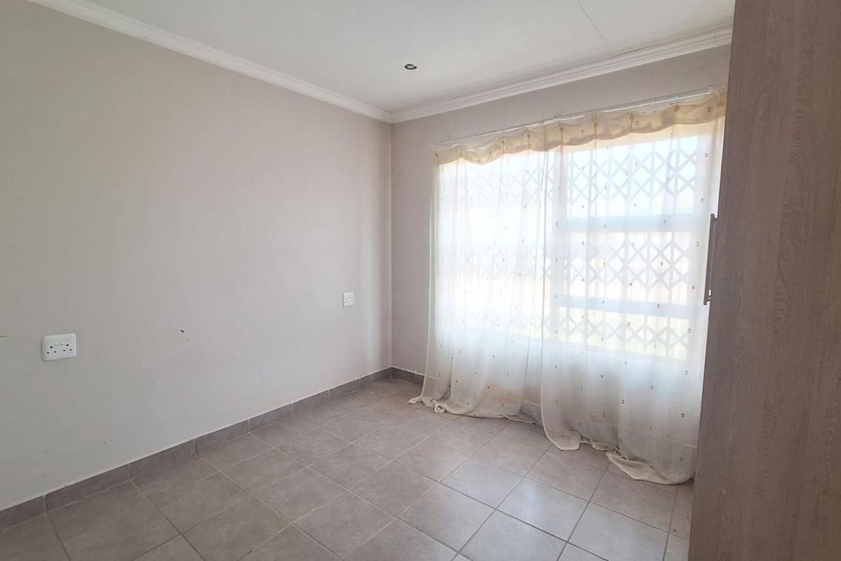 A bright, empty bedroom featuring tiled flooring, built-in cupboards, and a window fitted with a security gate.