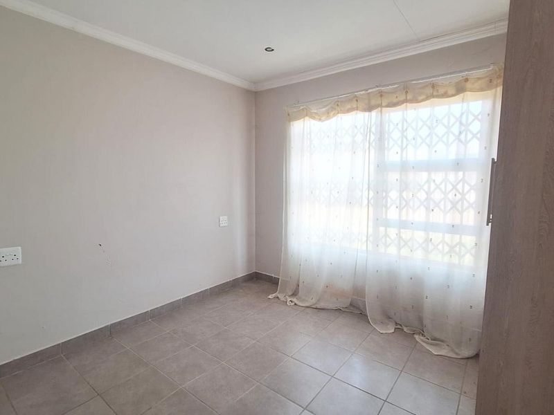 A bright, empty bedroom featuring tiled flooring, built-in cupboards, and a window fitted with a security gate.
