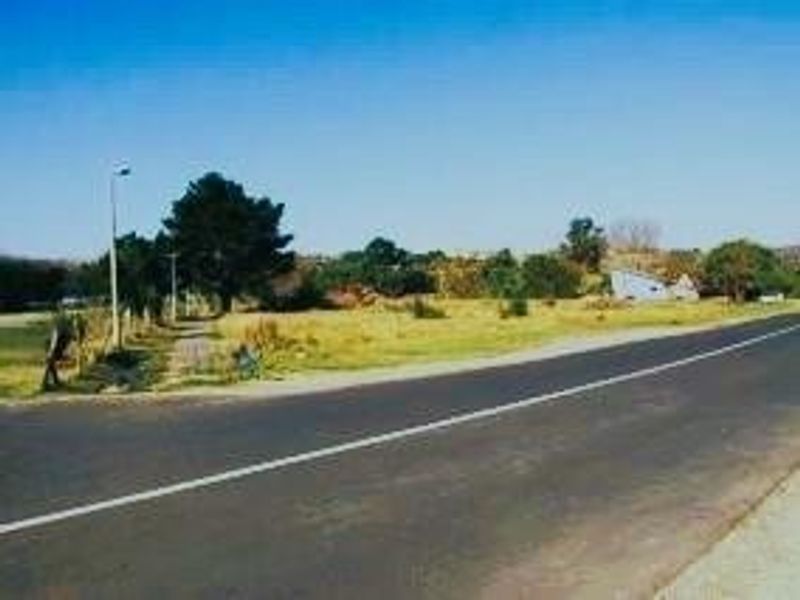 A street-level view of a vacant residential plot adjacent to an asphalt road with existing street lighting and perimeter fencing.