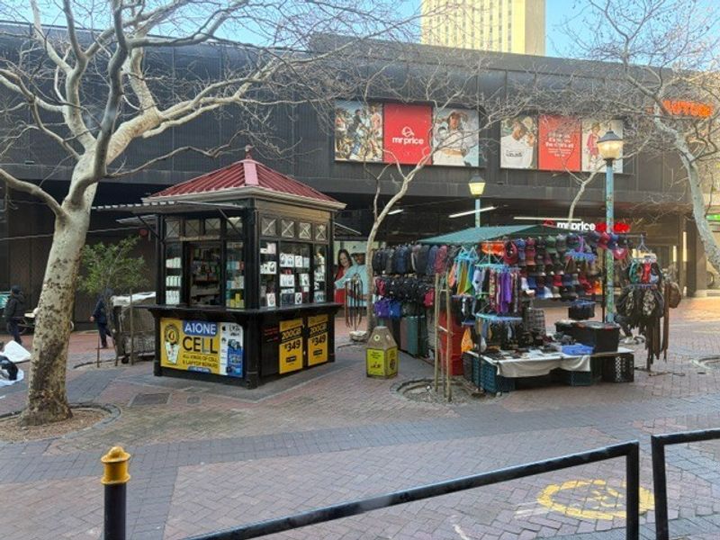 Exterior view of a retail shopping area featuring a permanent kiosk structure and paved pedestrian walkway in front of a commercial building facade.