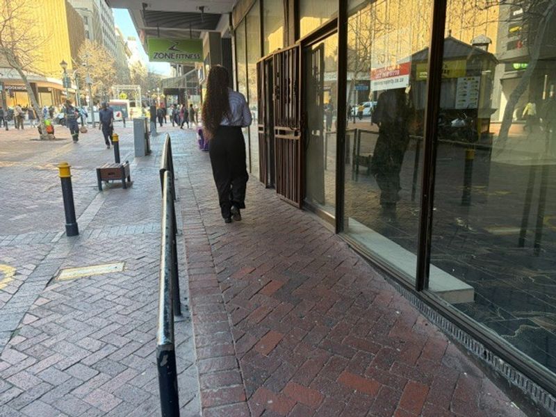 Street-level view of a retail storefront featuring large glass display windows and a metal security gate on a brick-paved pedestrian walkway.