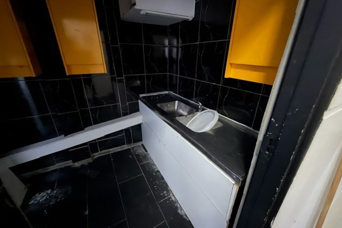 A small, dated kitchenette featuring black tiled walls, white base cabinetry, and a stainless steel sink, currently in need of refurbishment.