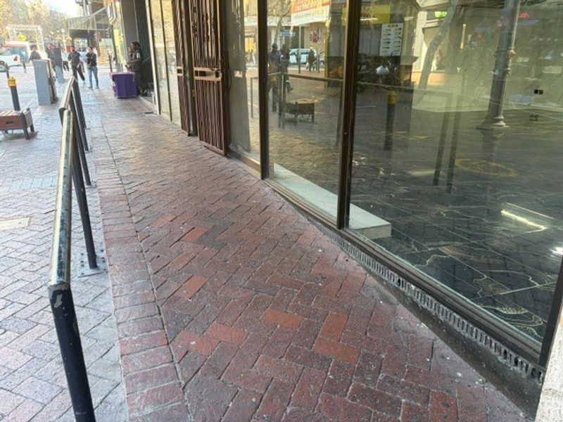 Street-level view of a retail storefront featuring large glass display windows, metal security gates, and brick-paved pedestrian access.