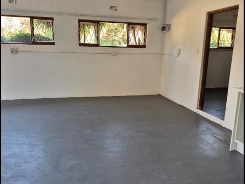 An empty living area featuring painted concrete flooring, security-barred windows, and exposed electrical conduit, offering a blank canvas for renovation.