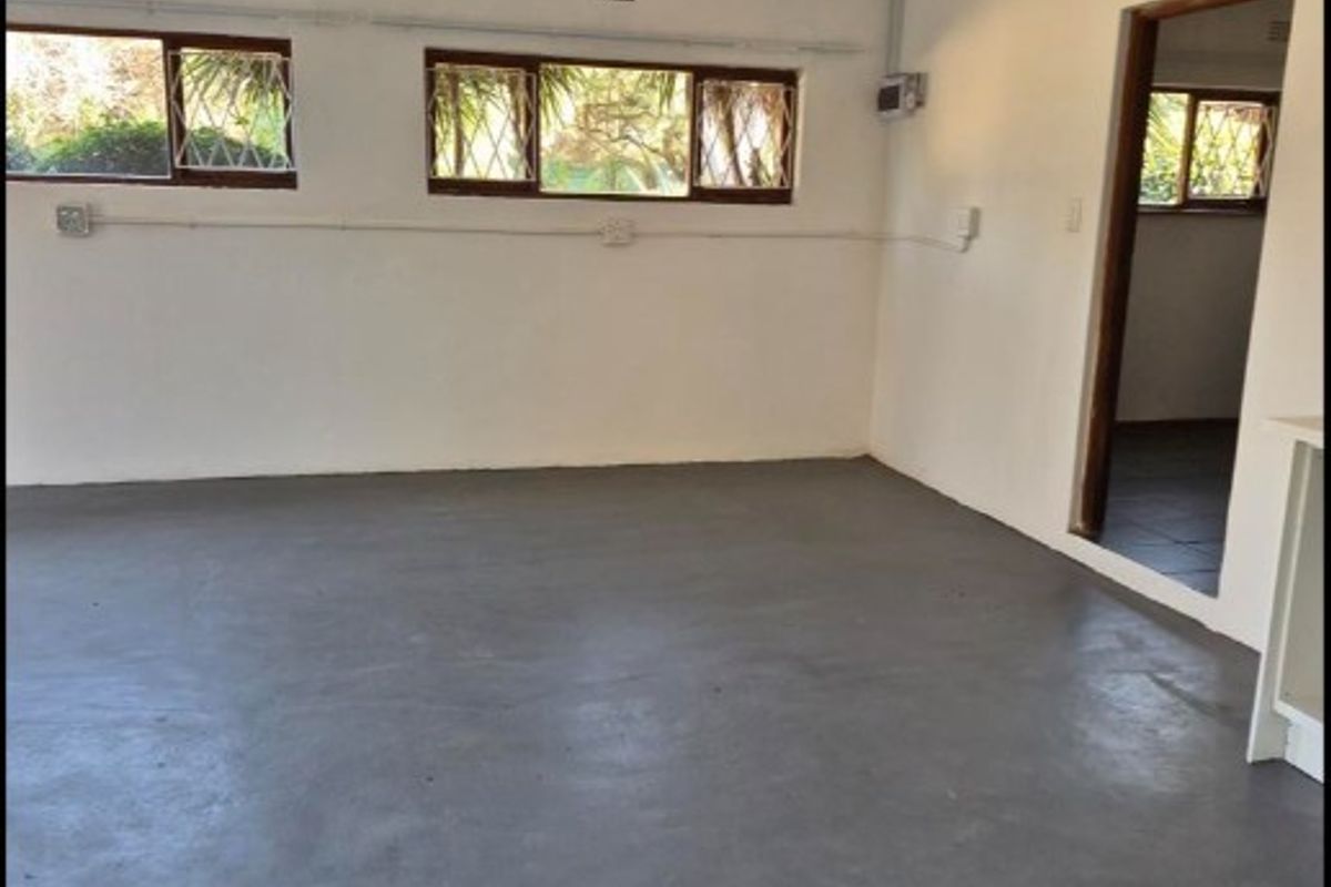 An empty living area featuring painted concrete flooring, security-barred windows, and exposed electrical conduit, offering a blank canvas for renovation.