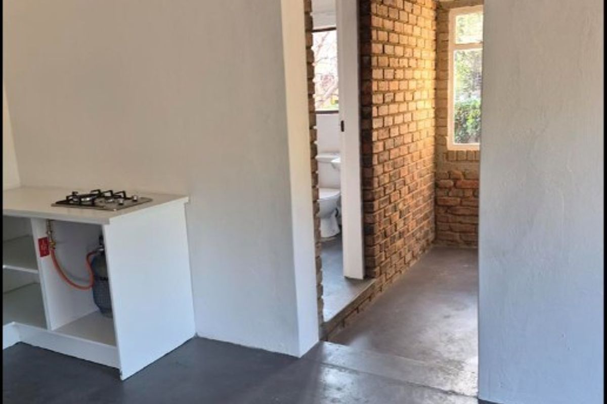 Open plan area featuring exposed brickwork, painted concrete floors, and a basic kitchenette with a gas hob, leading to an adjacent bathroom.