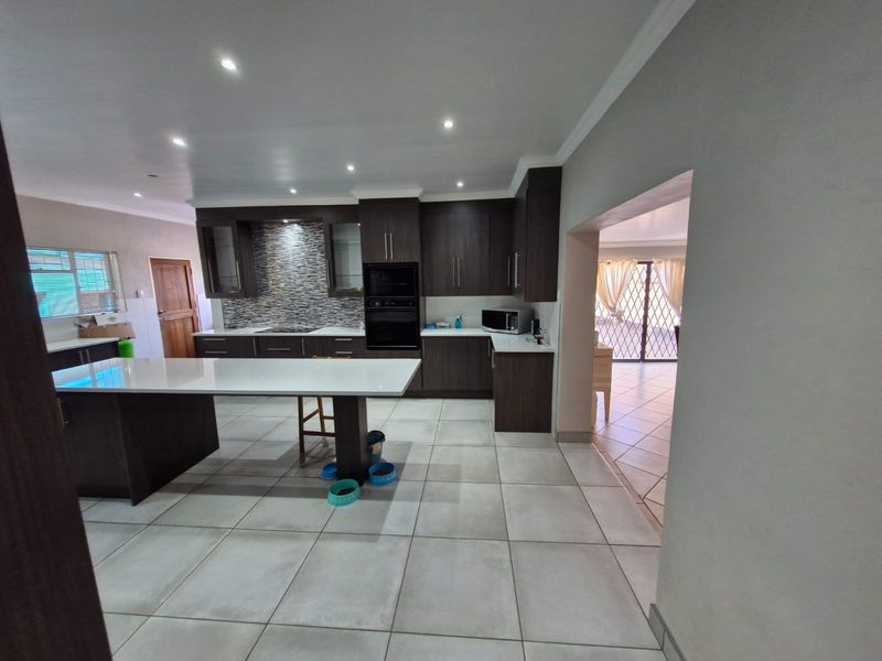 Modern kitchen featuring a large central island, dark cabinetry, stone countertops, mosaic backsplash, and integrated appliances with neutral tiled flooring.