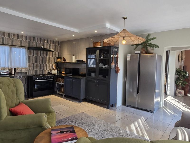 An open-plan kitchen and living area featuring modern dark cabinetry, a tiled backsplash, and neutral floor tiles with recessed ceiling lighting.