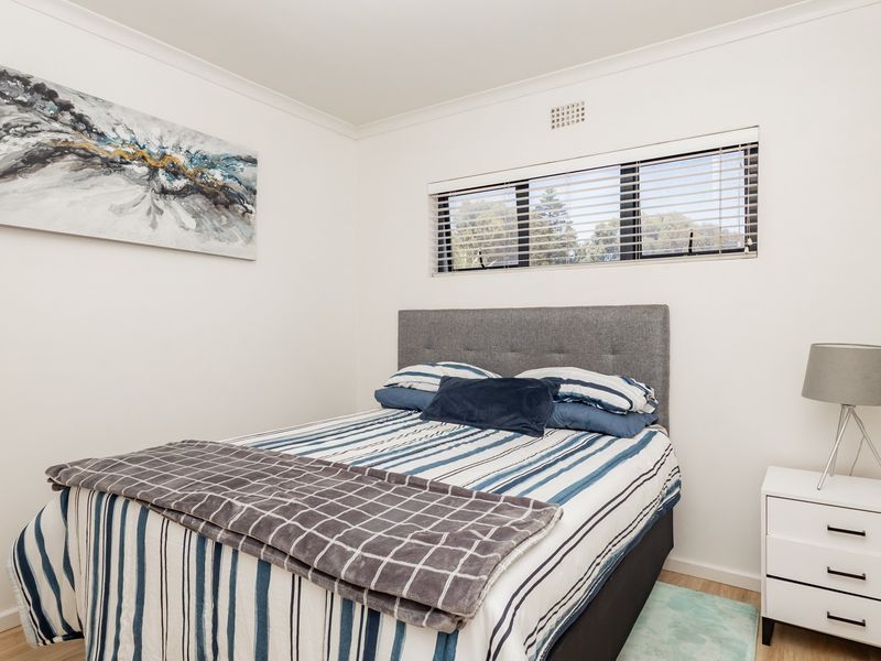 A well-maintained bedroom featuring neutral walls, laminate flooring, and a window fitted with venetian blinds and a ventilation grille.