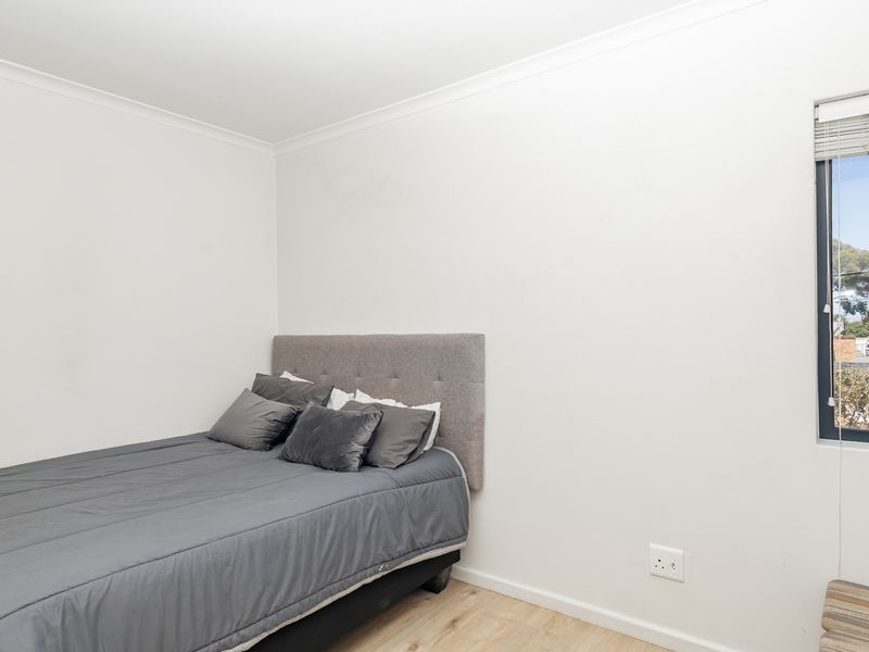 A bright bedroom featuring neutral walls, laminate flooring, and a window with venetian blinds, offering a clean and functional living space.