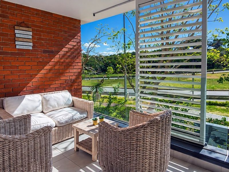 A covered balcony featuring an exposed brick wall, glass balustrade, and adjustable louvered shutters, offering a pleasant outdoor seating area with scenic views.