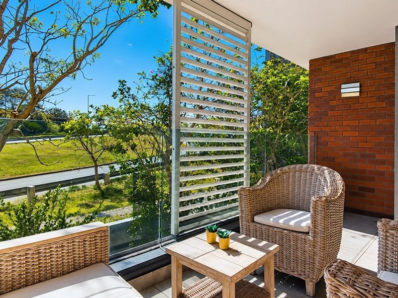 A modern balcony featuring a glass balustrade, adjustable louvred privacy screen, and an exposed brick feature wall, overlooking a green landscape.
