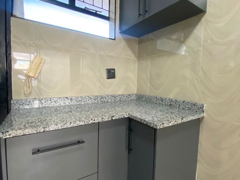 A compact kitchen area featuring modern grey cabinetry, granite countertops, and neutral tiled walls with security bars on the window.