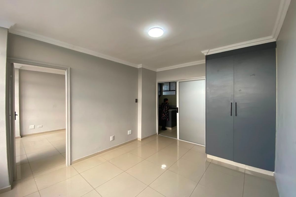A bright, empty living area featuring neutral tiled flooring, coving, and a modern built-in cupboard, with access to an adjacent room and kitchen area.