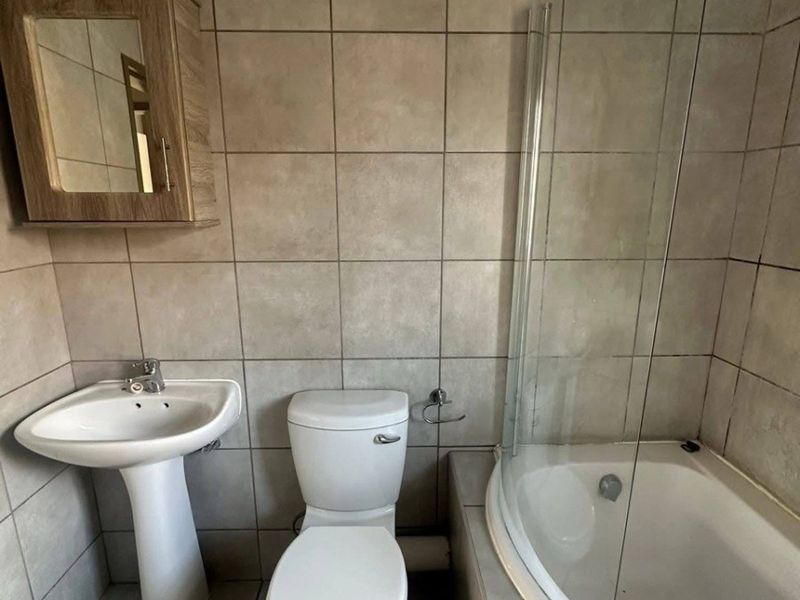 A functional bathroom featuring a pedestal basin, toilet, and a bathtub with a glass shower screen, finished with neutral wall and floor tiles.