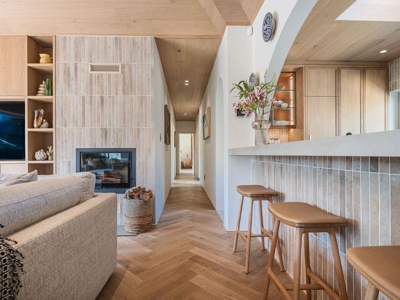 Modern open-plan living area featuring herringbone timber floors, a tiled fireplace feature wall, and a stone breakfast bar connecting to the kitchen.