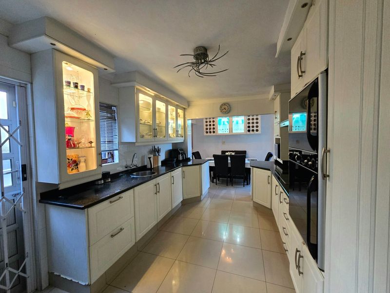 A well-maintained kitchen featuring white cabinetry, granite countertops, built-in oven, and glass-front display units, leading into an adjacent dining area.