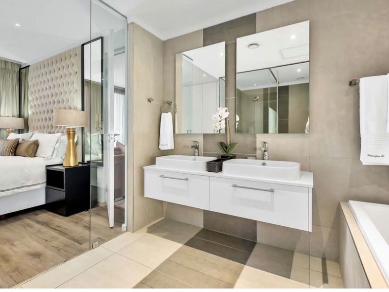 Modern en-suite bathroom featuring a sleek double vanity with vessel sinks, large mirrors, and contemporary tiled finishes, seamlessly connected to the bedroom via glass partition.