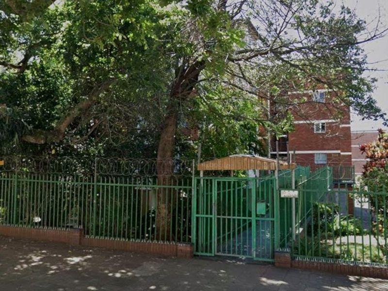 Front exterior view of the apartment complex featuring brick facade, metal perimeter fencing with security razor wire, and a pedestrian entrance gate.
