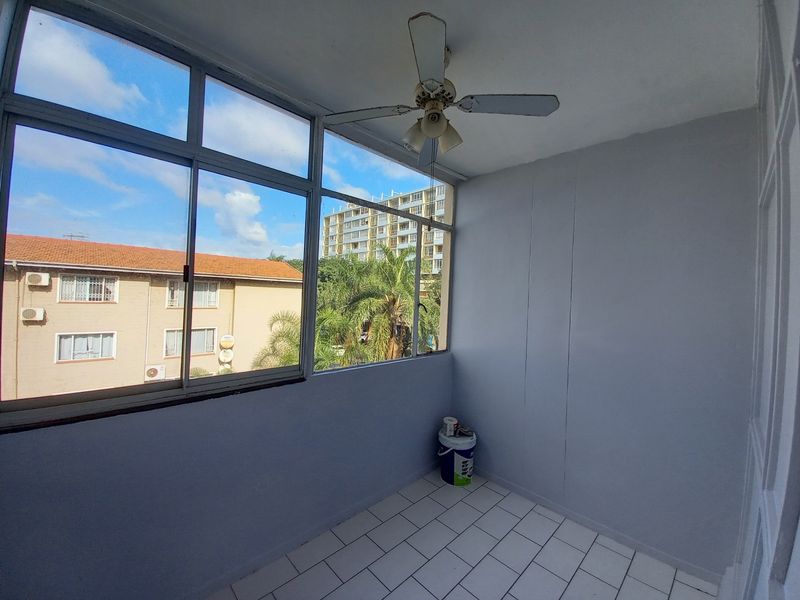 An enclosed balcony featuring tiled flooring, a ceiling fan, and large windows offering an urban view, suitable for a sunroom or study area.