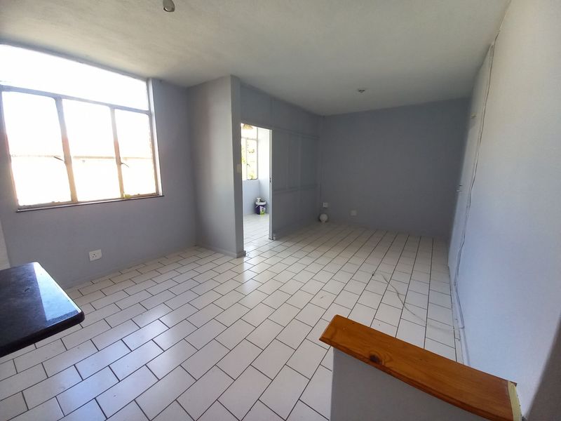 An empty open-plan living area featuring white tiled flooring, neutral grey walls, and a large window providing natural light.